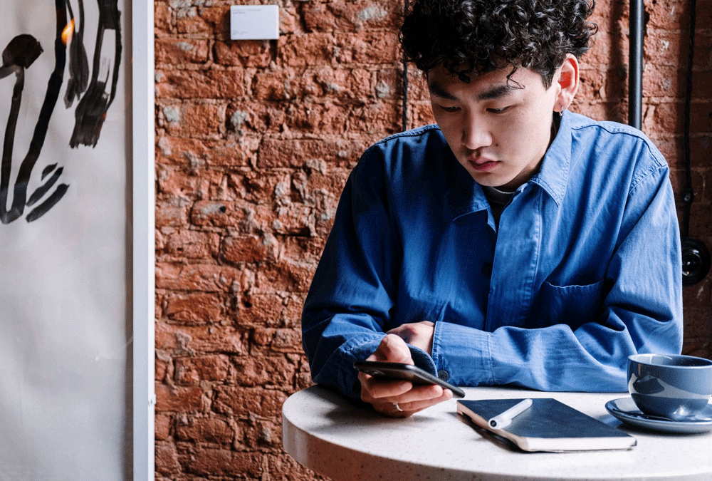 A millennial sits at a round coffee table on his phone at a table with coffee and a notebook. There is a brick background behind him.