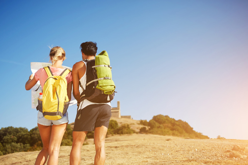 A woman and man with backpacks stop to look at a map on a dry hillside. You cannot see their faces and they are dressed sporty.