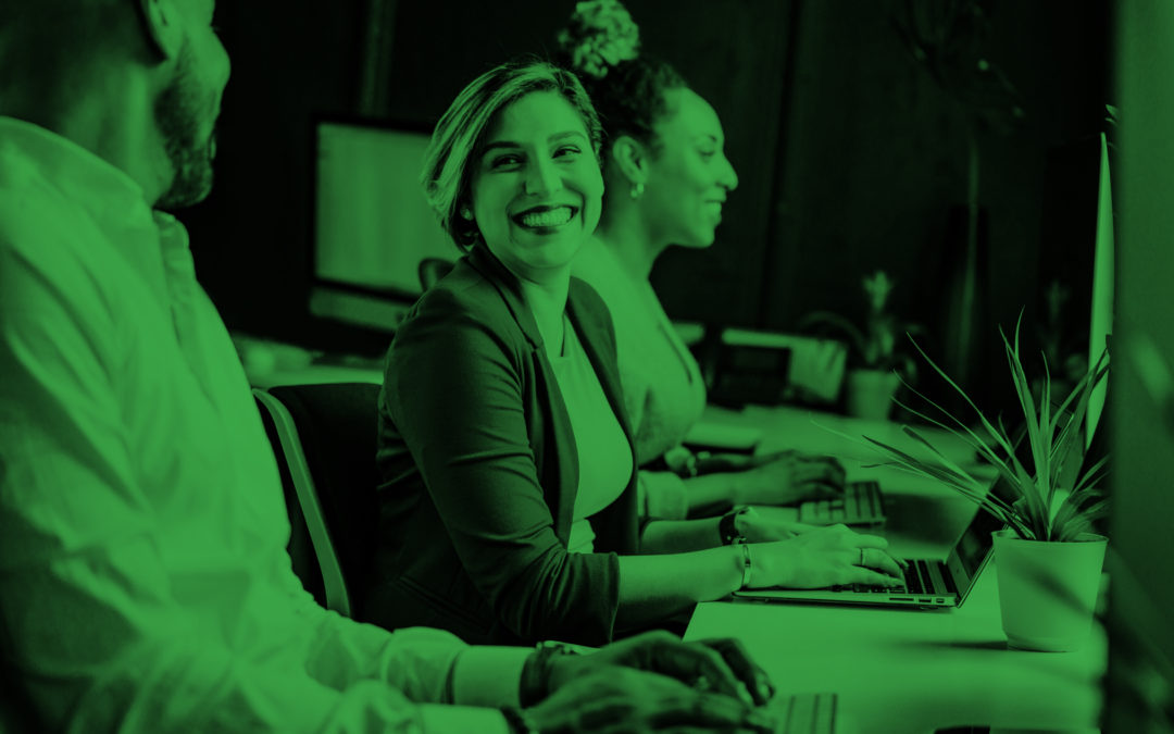 Three coworkers sit at their desk on a computer. Two coworkers smile at each other.