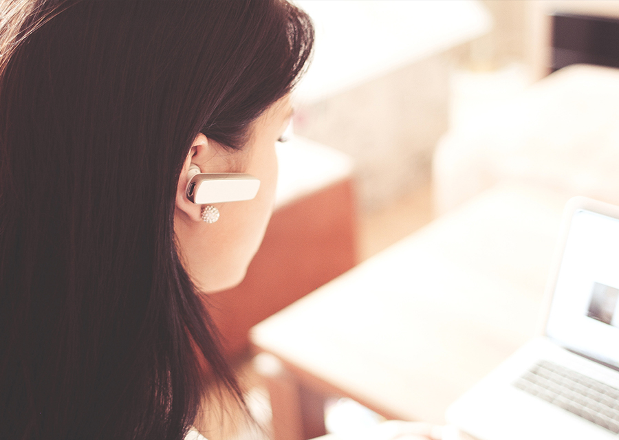 A woman wearing bluetooth headphones reads from her computer. The laptop rests on a wooden coffee table.