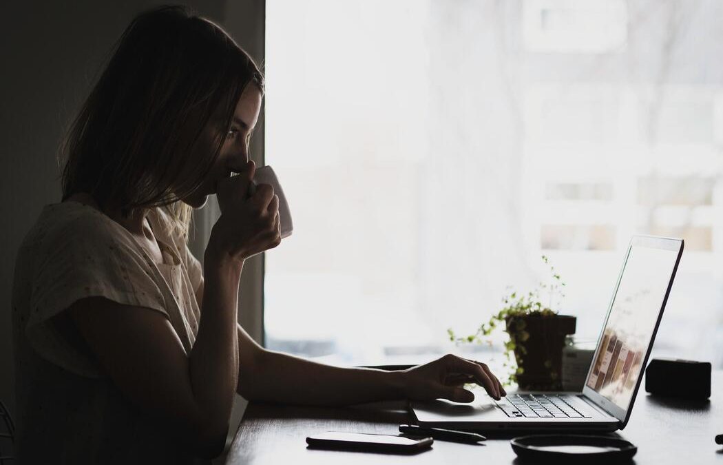 A woman sips from a coffee mug while working on her laptop.