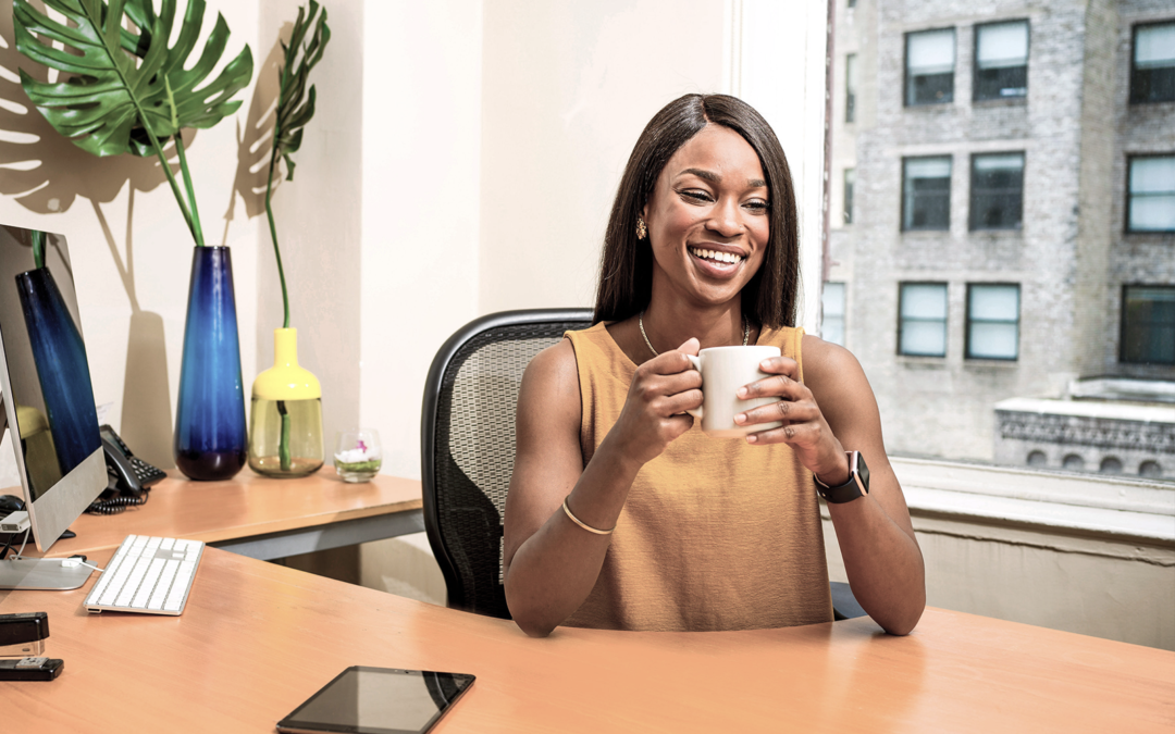 A woman sits at her desk smiling with a cup of coffee in hand. The desk has fake plants and multiple devices on it.