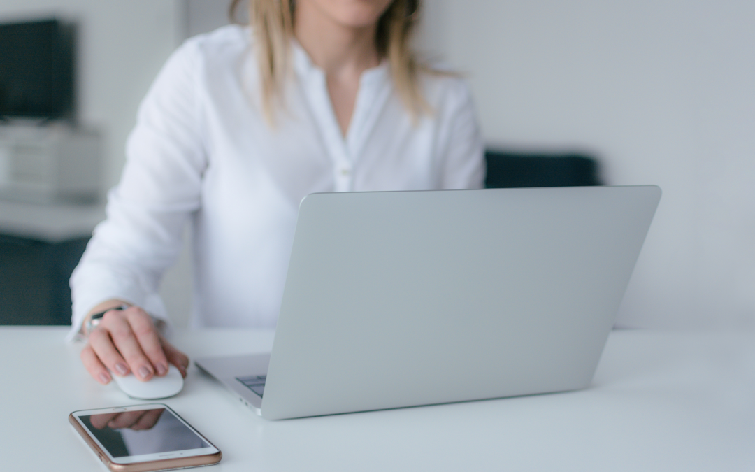 A woman clicks on her computer mouse. A phone lays next to the open laptop. You can only see her lips and torso.