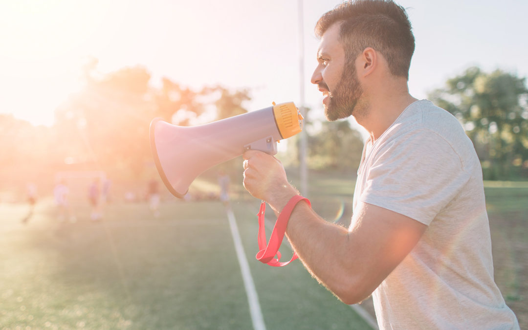 A male soccer coach holds a megaphone to his face. You can see his torso and the side profile of his face. He is on a turf field.