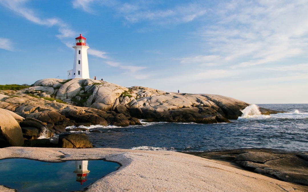 A white lighthouse next to the ocean. Waves crash into the rock cliff and people stand looking at the view.