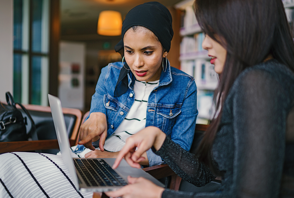 Two woman sit at chairs in a library. They are both pointing at a laptop screen.