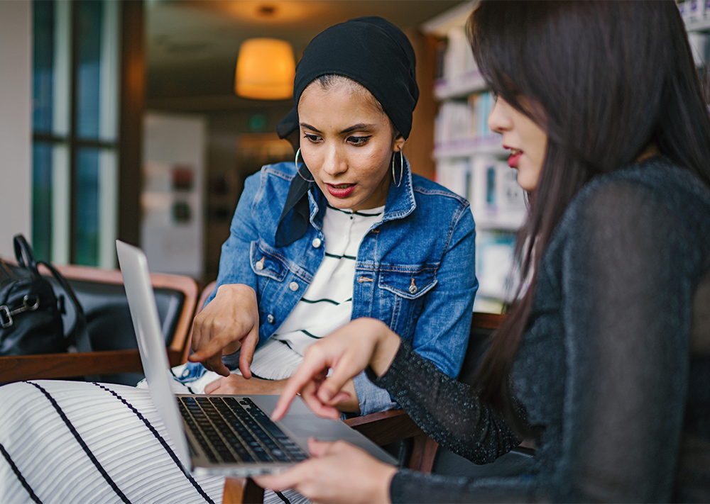Two woman sit at chairs in a library. They are both pointing at a laptop screen.