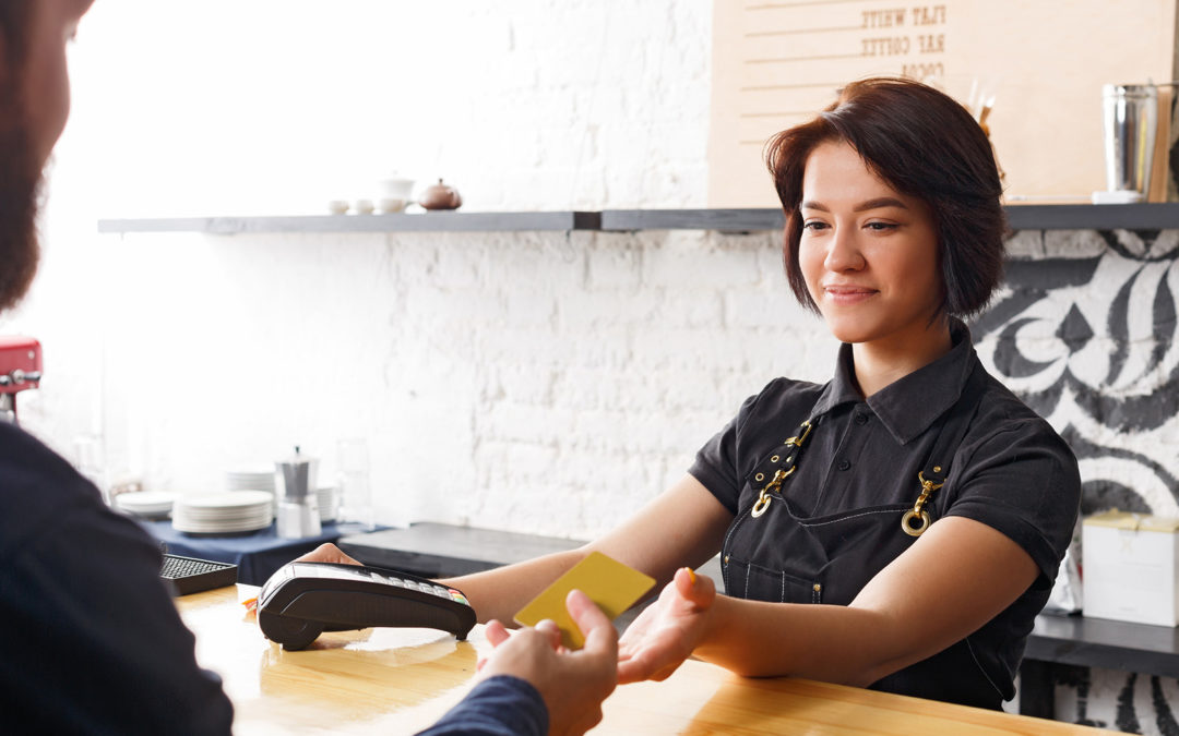 A barista accepts card payment from a business man. You cannot see the mans face, only the barista's, who is a woman wearing an apron.