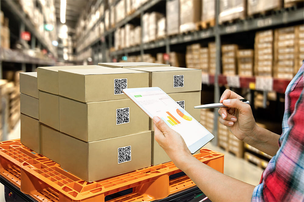 A person holding a tablet in front of a pallet of manufactured goods.