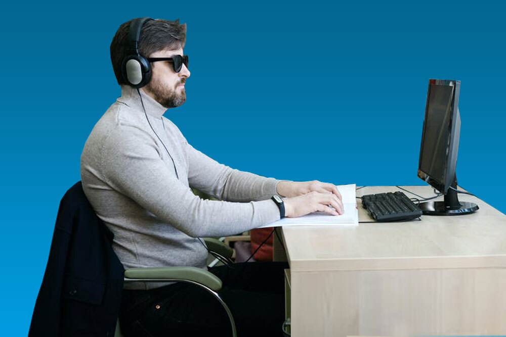 A picture of a man reading braille in front of his computer. He has sunglasses and headphones on.