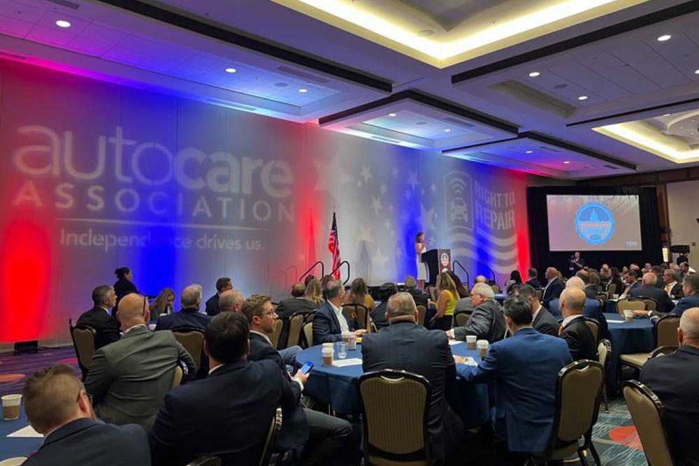 Image of auto care conference. Multiple people sitting and listening to a speaker on stage.